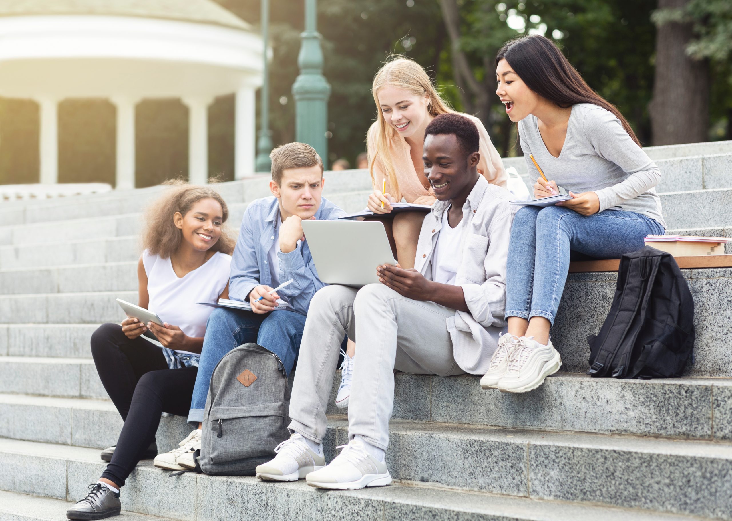 Internationale Studierende, die auf einer Treppe im Park sitzen. Sie schauen gemeinsam auf einen Laptop und lachen dabei.